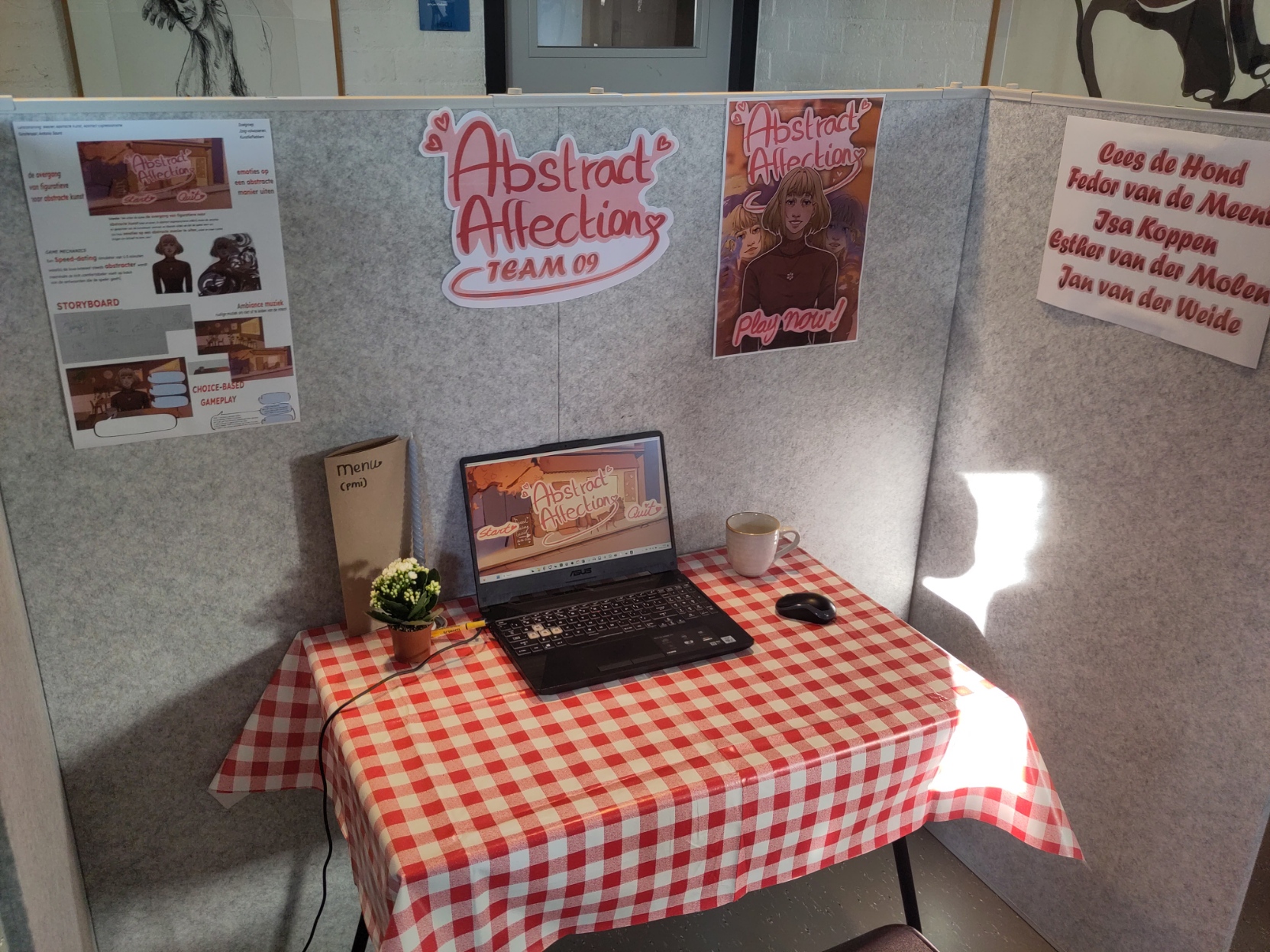 A small dining table with a plant, coffe mug, menu and a gaming laptop with a game called "Abstract Affection.

There are gray walls around the table with a poster, logo and a document explaining game design.

The logo says "Abstract Affection Tram 09".

There is also a credits page with the names "Cees de Hond", "Fedor van de Meent", " Isa Koppen", "Esther van der Molen" and "Jan van der Weide"