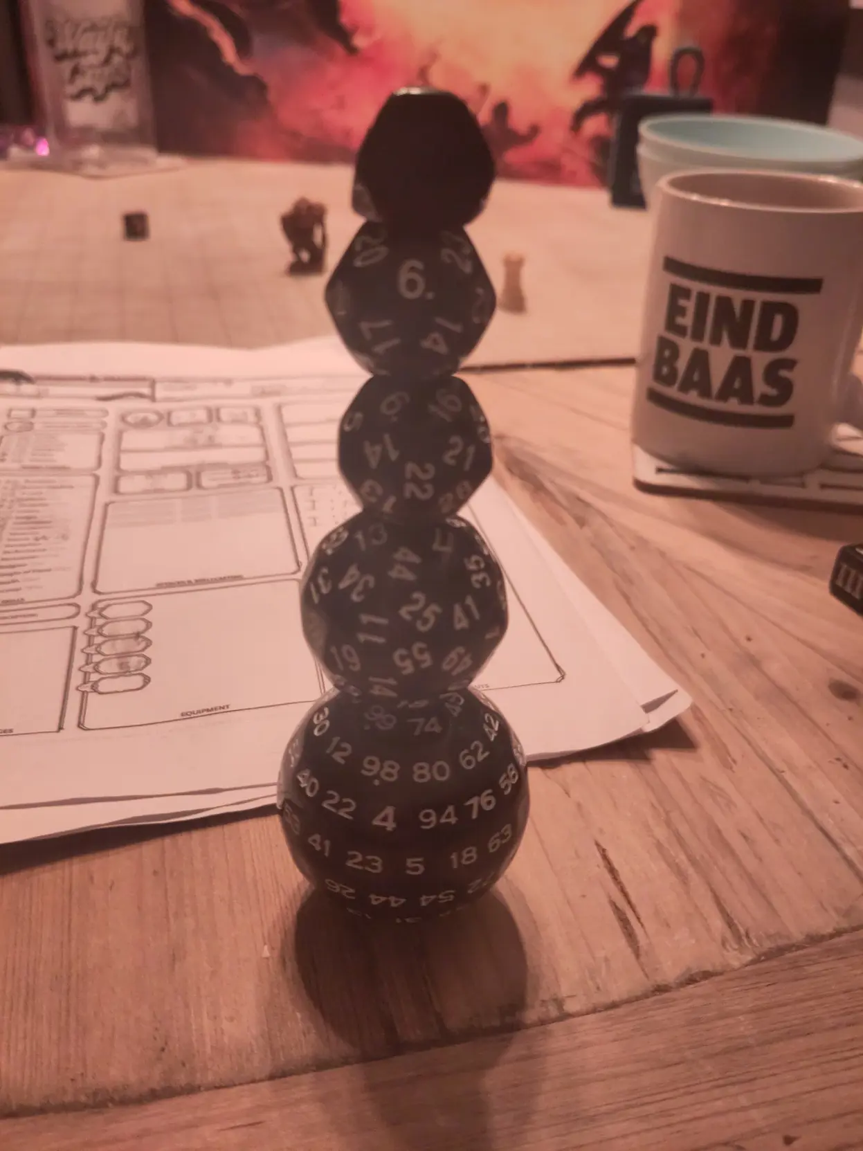 A stack of dice (a d100, d60, d30 and a d24).

With a table filled with dnd stuff and mugs.